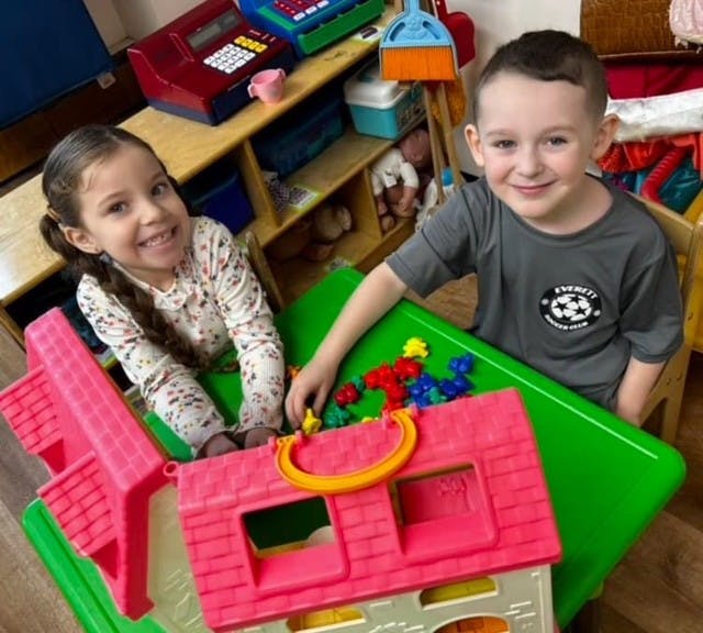 Two children playing with a toy house and plastic figures at a table