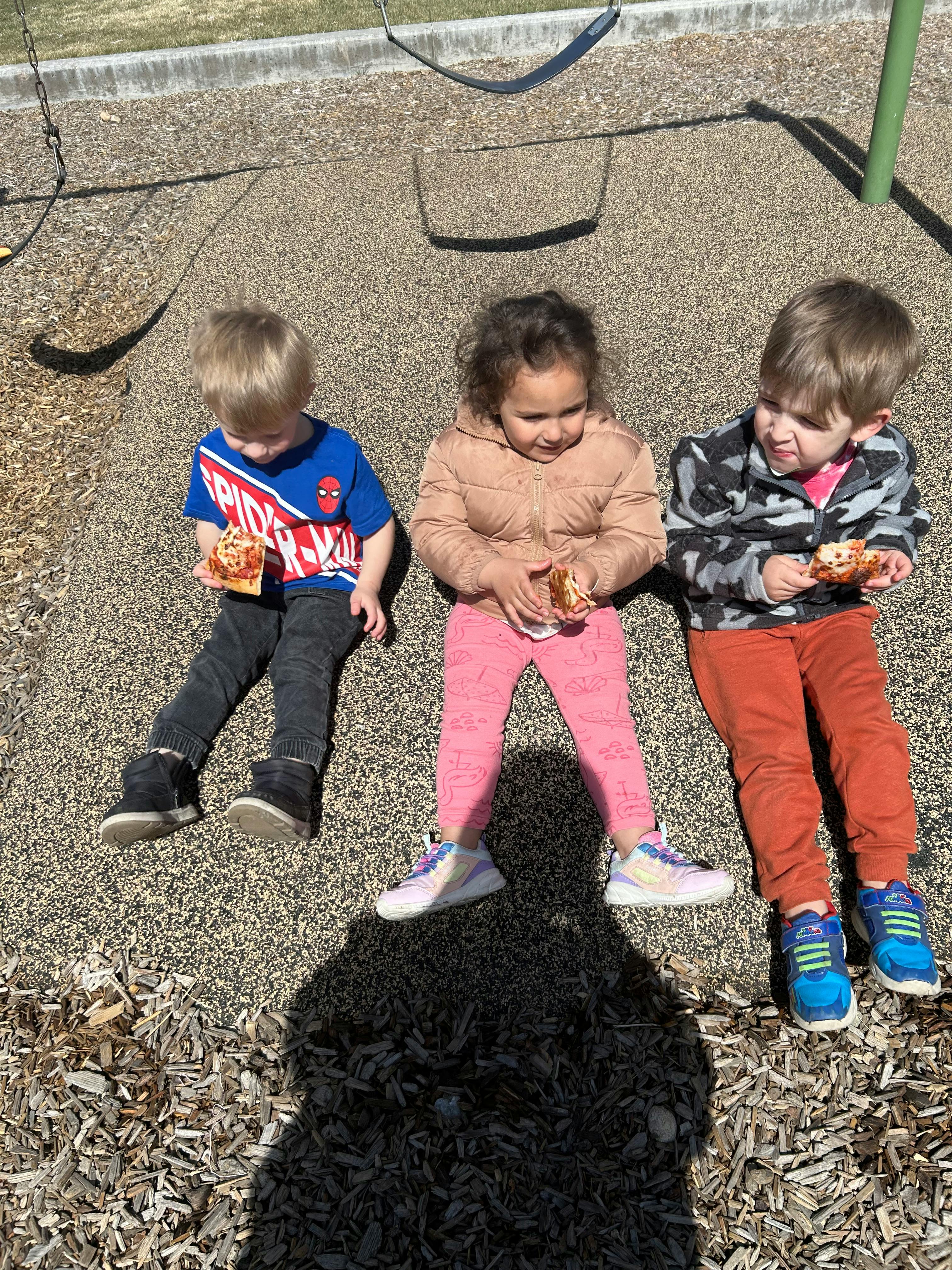 Three children sitting on wood chips eating pizza on a playground