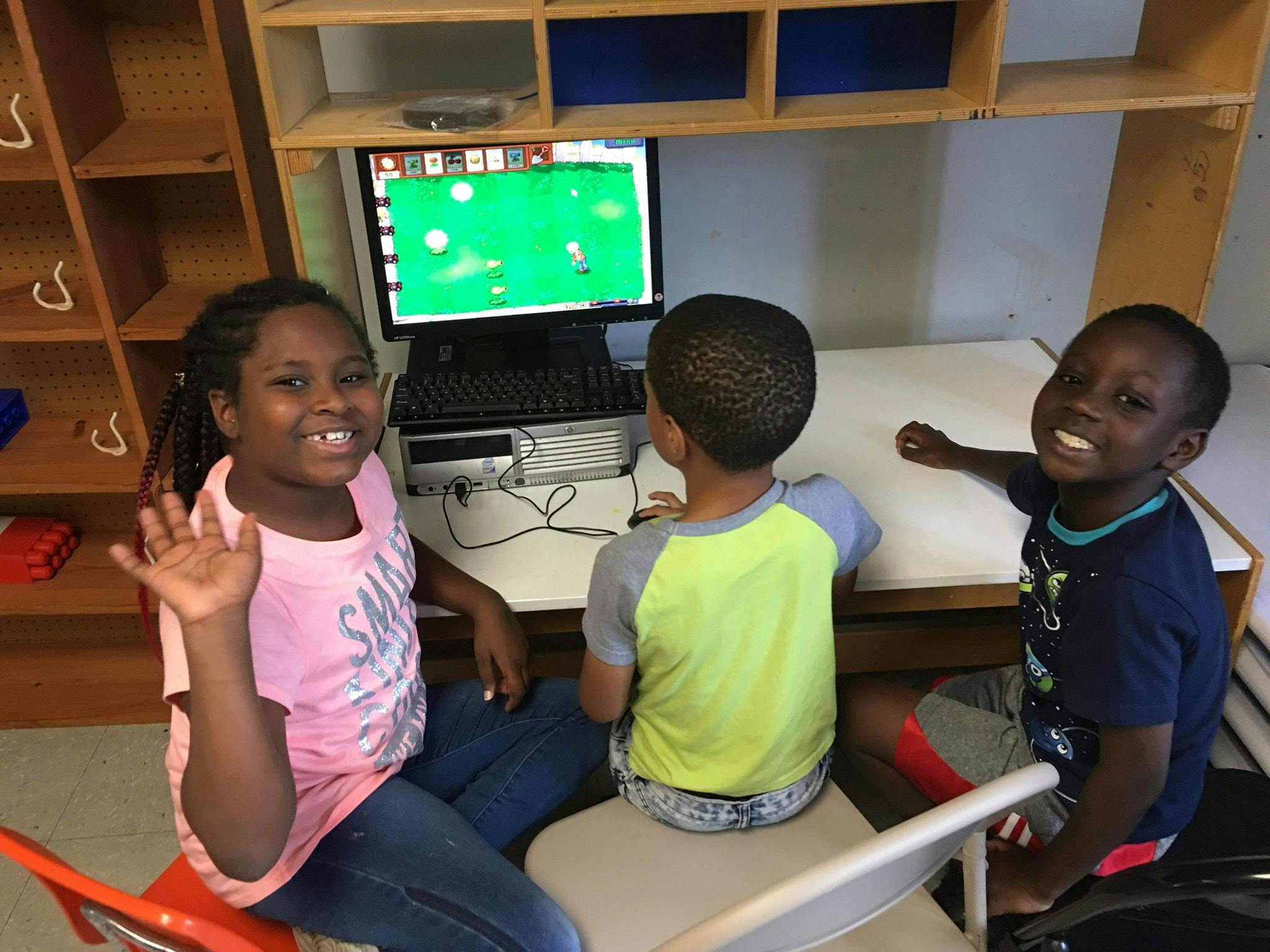 Three children at a computer desk, one waving
