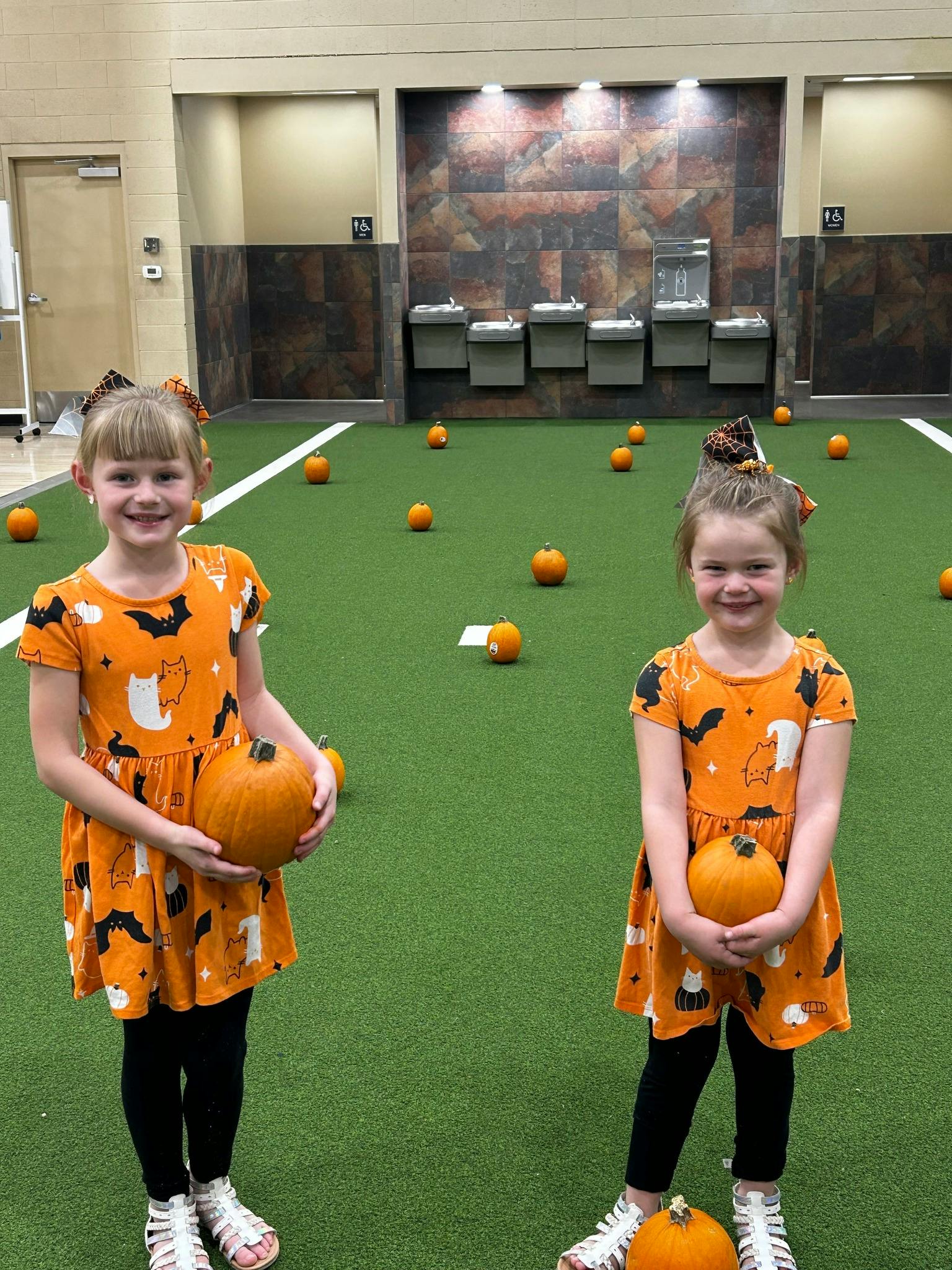 Two children in orange dresses holding pumpkins on a green turf field