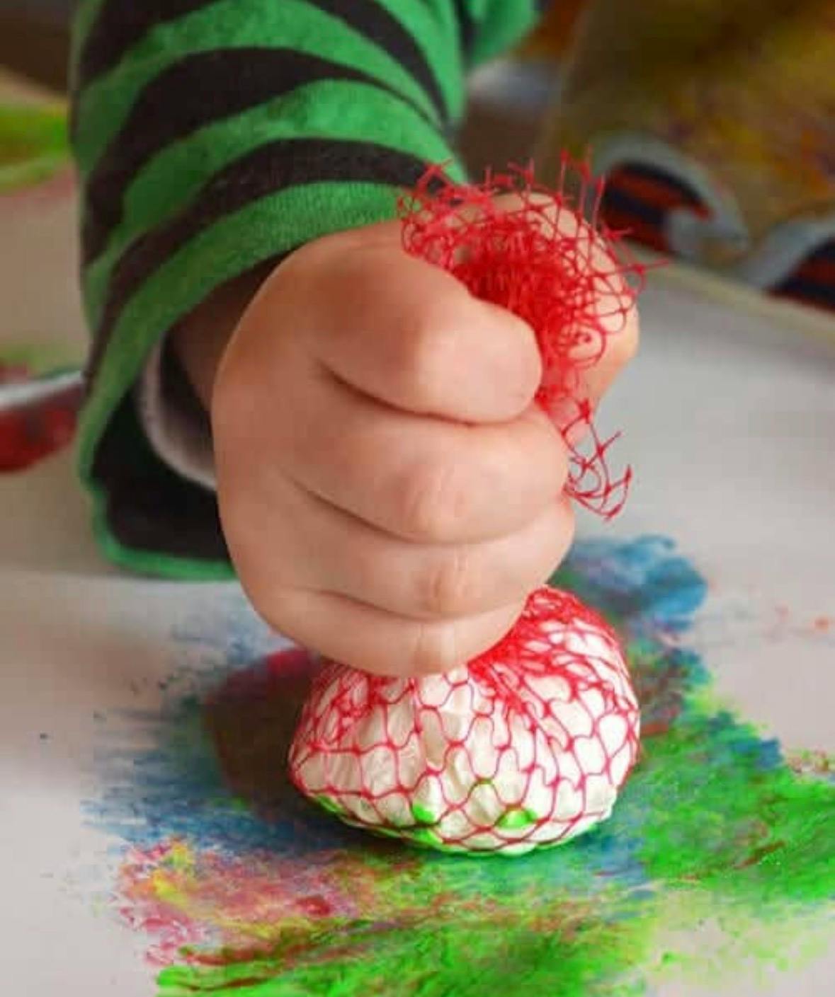 Child hand pressing a net-wrapped object into colorful paint on paper