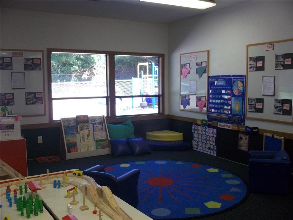 Classroom corner with windows, a bookshelf, blue rug, and educational displays
