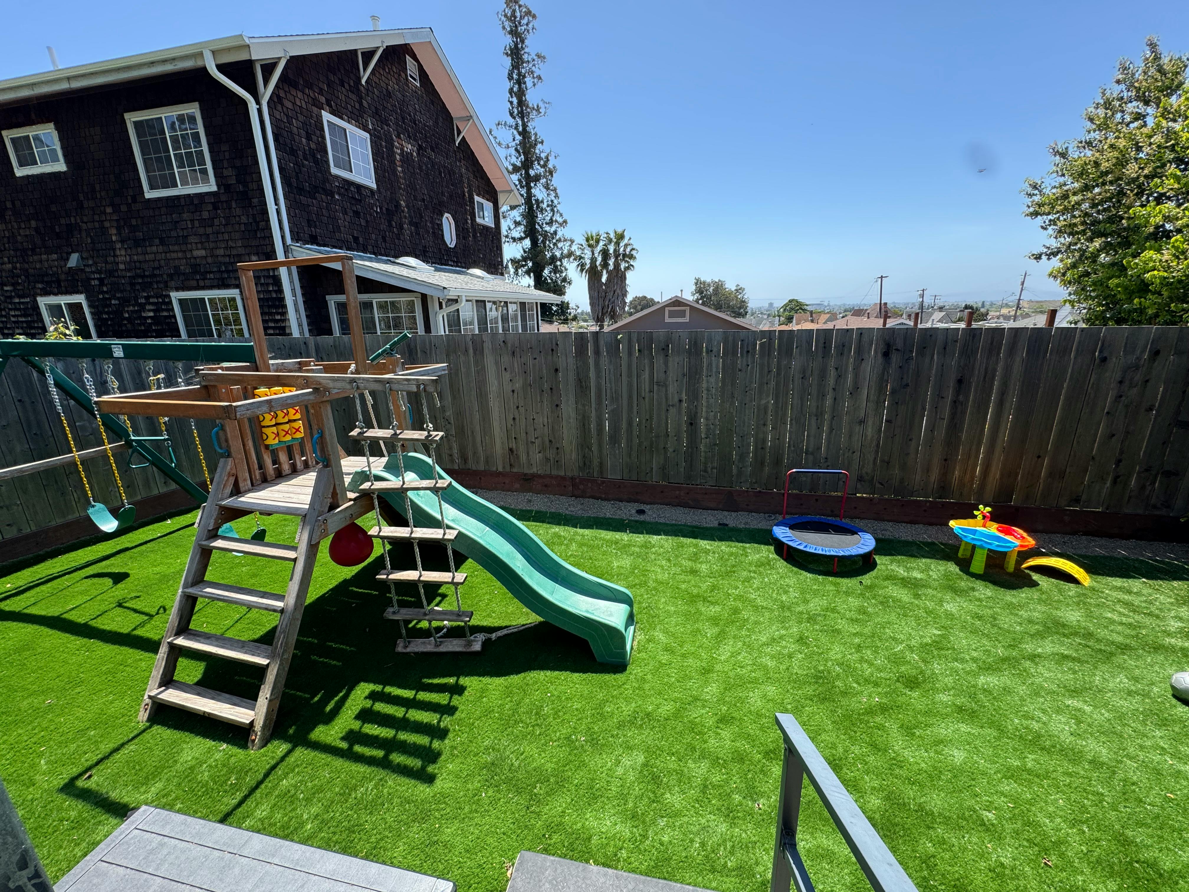 Backyard with a wooden playset, slide, and swing set on artificial turf