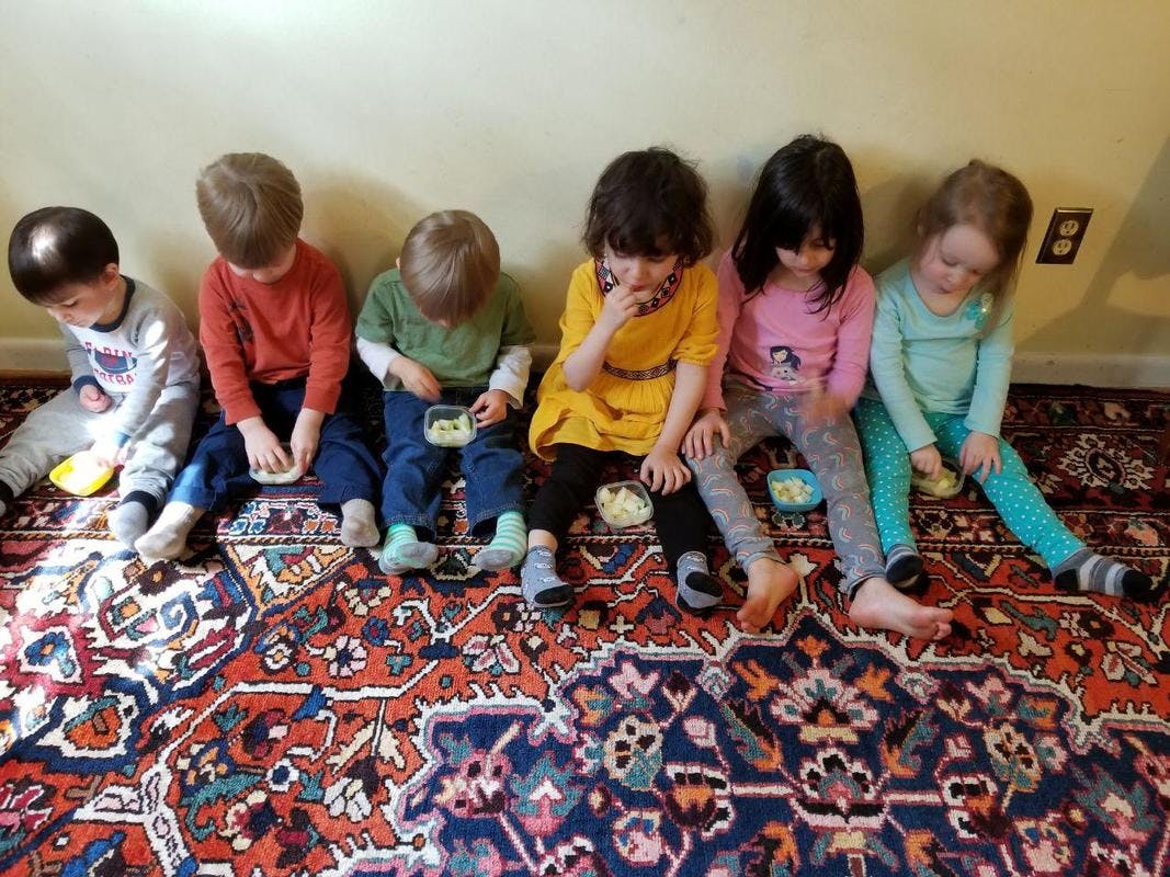 Six children sitting on a rug eating snacks in a row, eating snacks from small bowls
