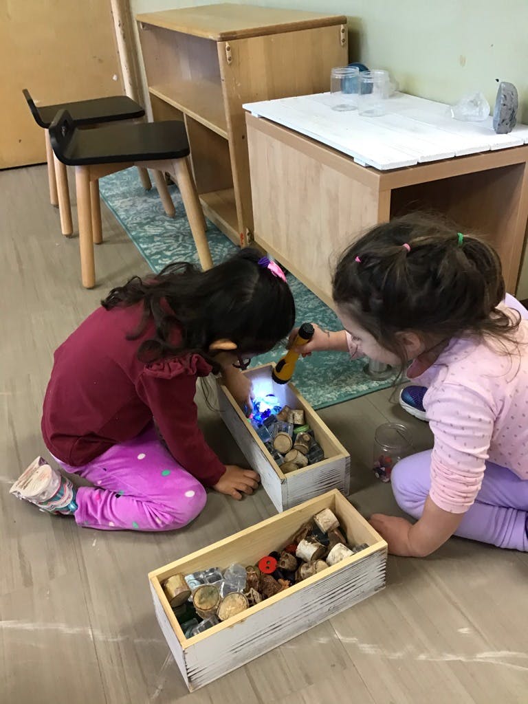 Two children on the floor looking at items in wooden boxes, one holding a flashlight