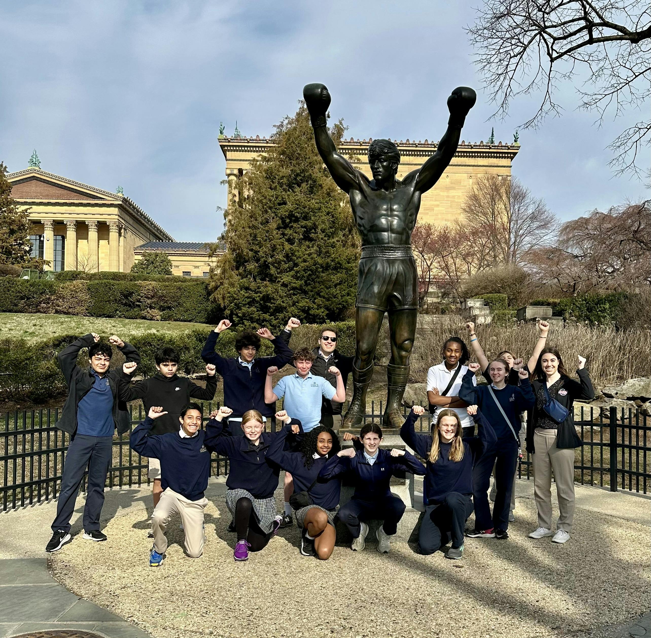Group of teens posing with a large statue of a person with arms raised