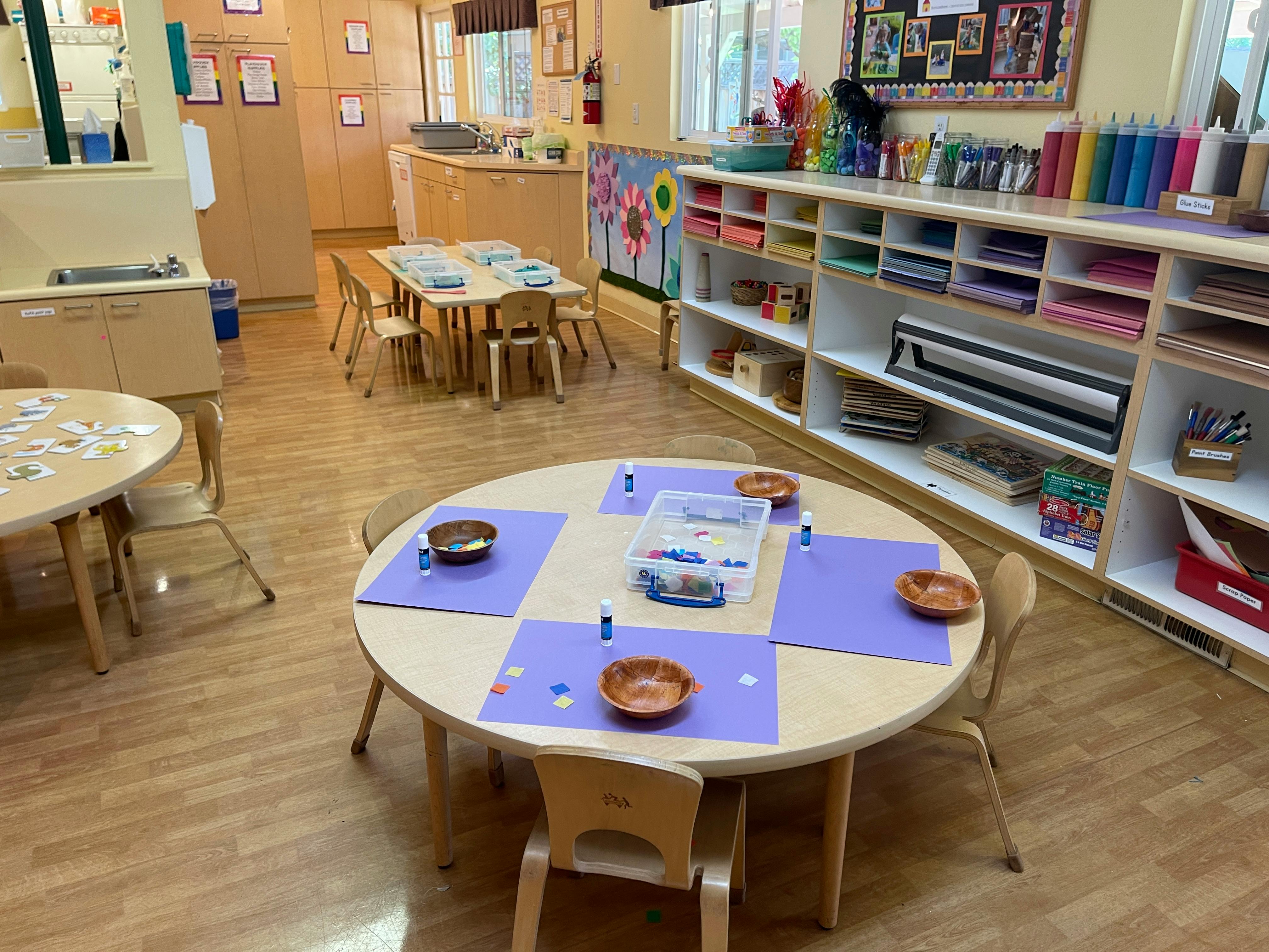 Classroom with small tables and chairs, art supplies on shelves