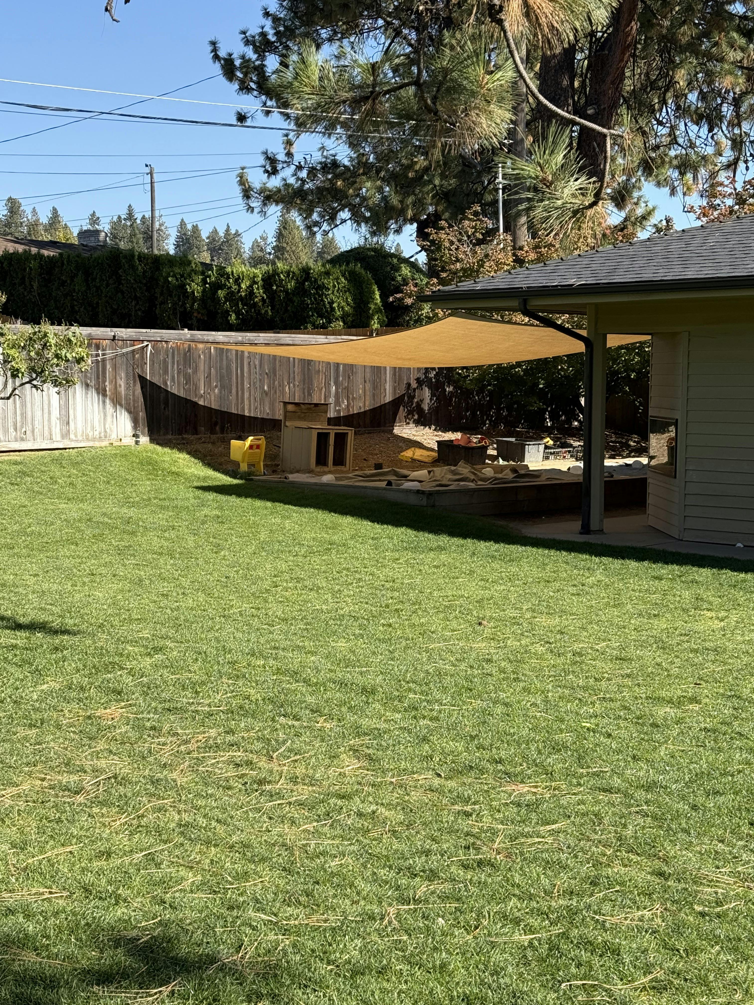 backyard with a wooden fence, green grass, and sandpit shaded by a tarp