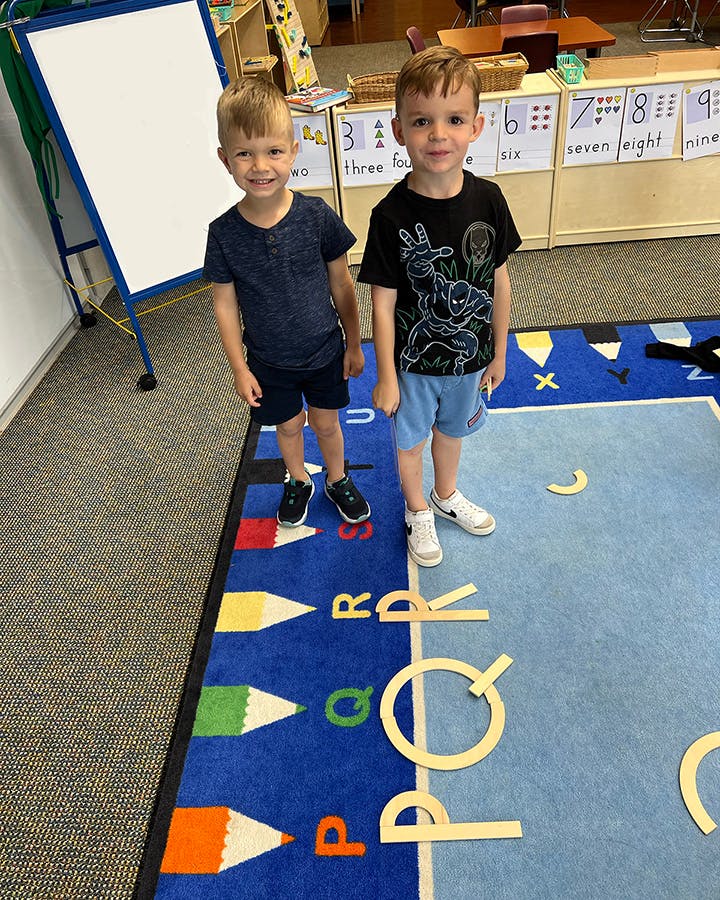 Two children standing on a rug with letters in a classroom