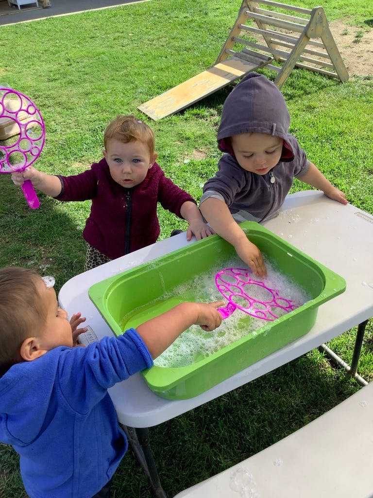 Three children playing with bubbles in a green tub on a table outdoors