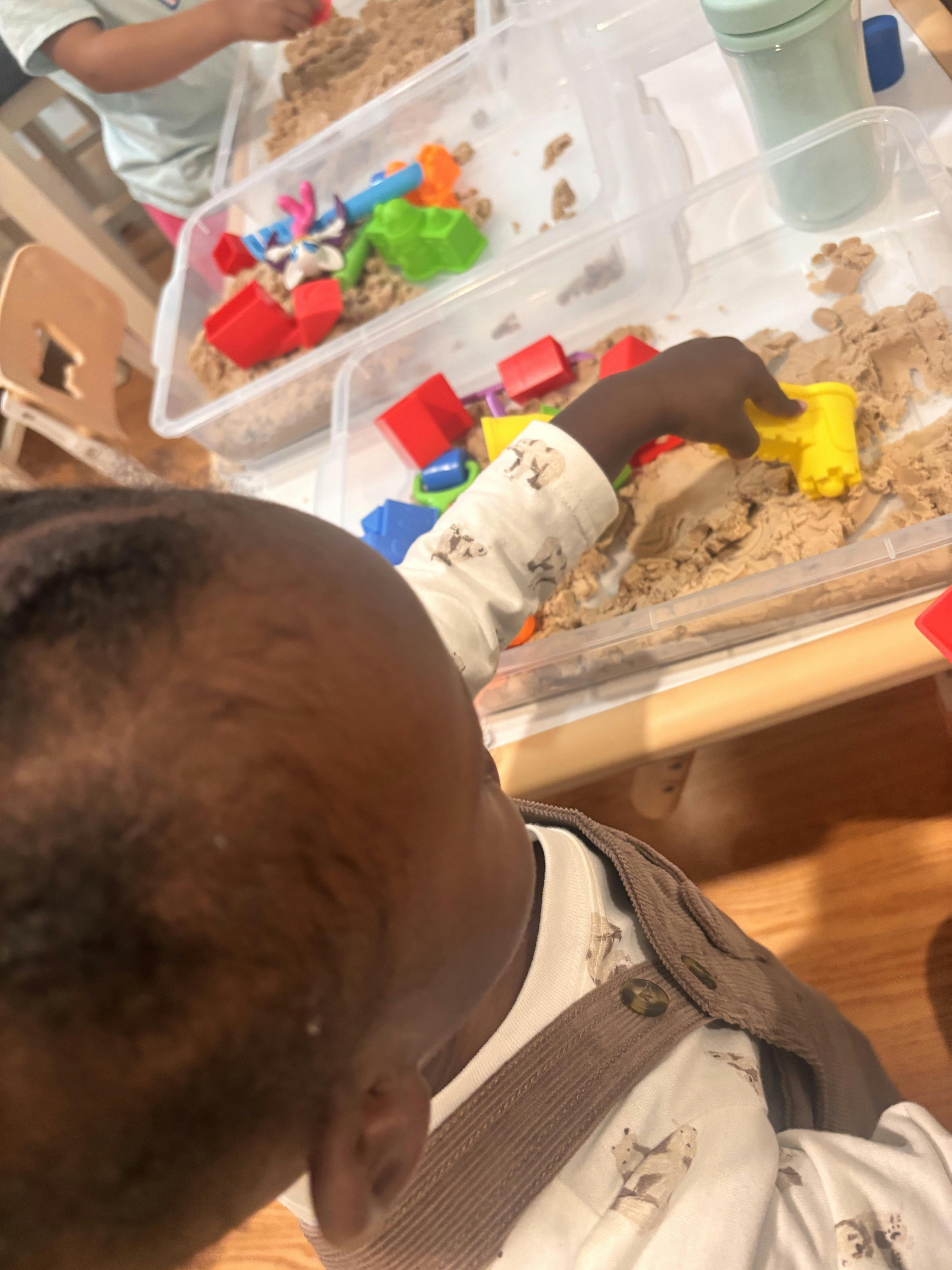 Child playing with kinetic sand and plastic molds in a clear storage bin
