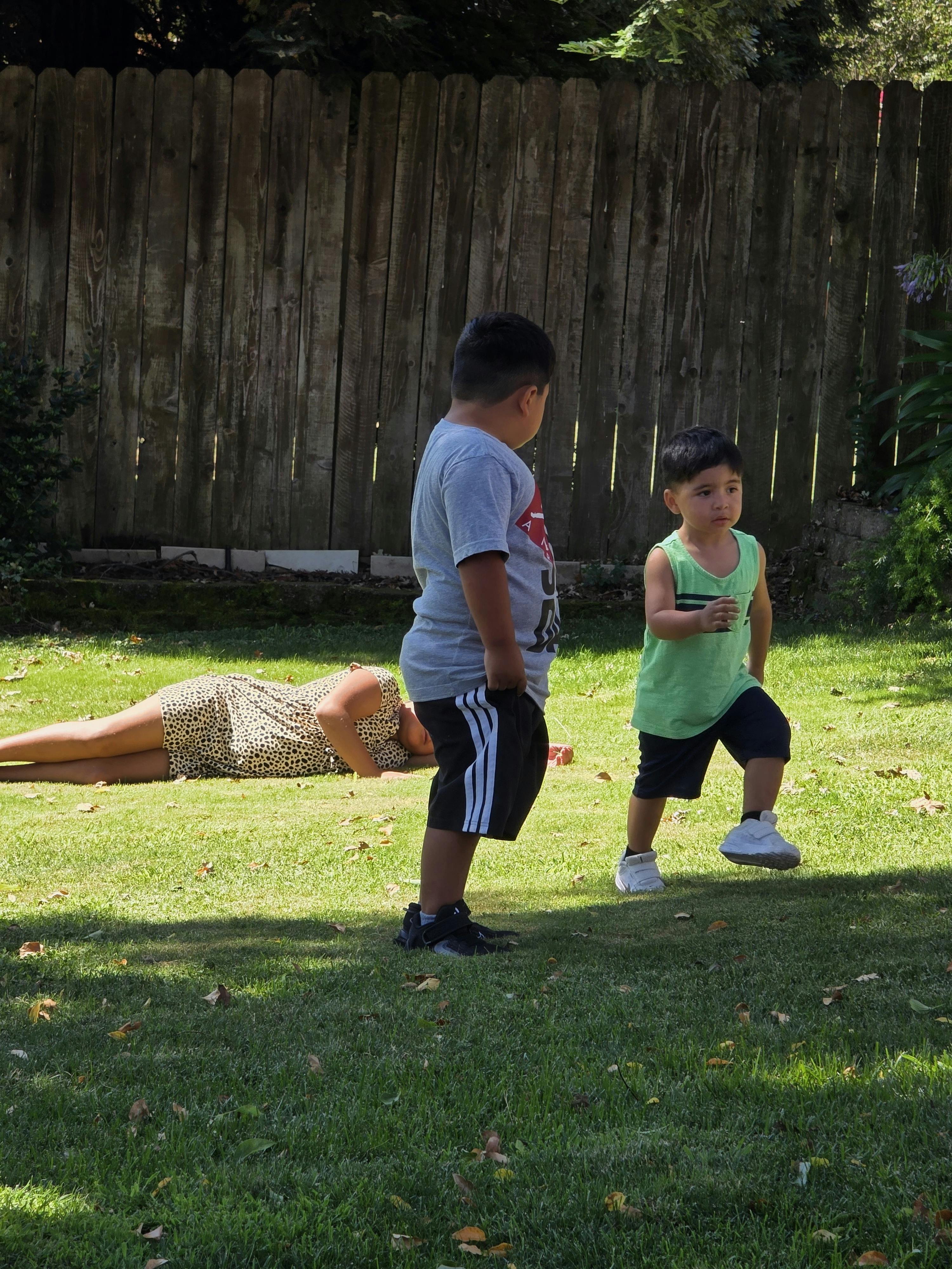 Two children standing on a lawn while another person lies on the grass