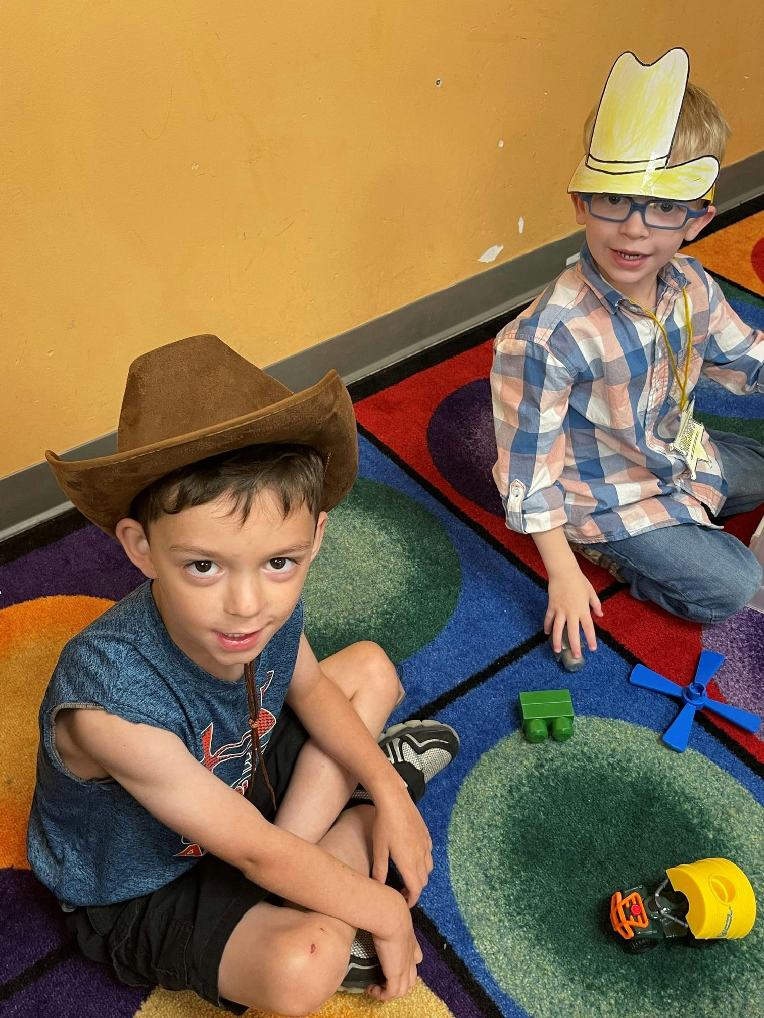 Two children sitting on a colorful rug, one wearing a cowboy hat