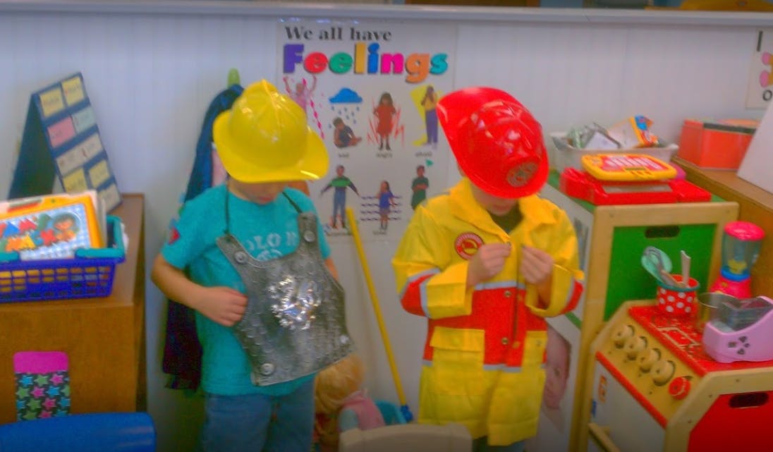 Two children in dress-up costumes, one in a yellow hard hat, one in a red firefighter helmet