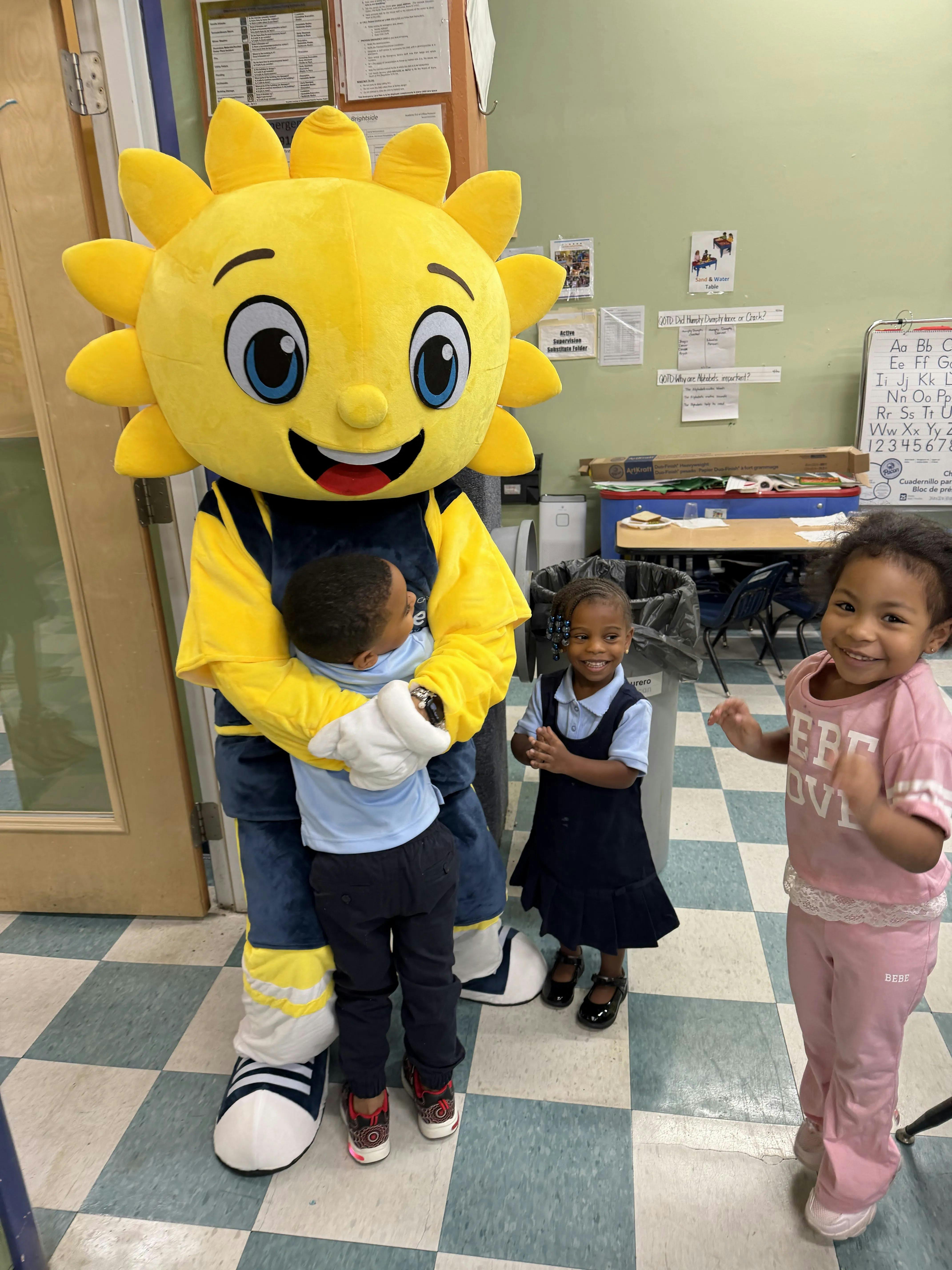 Child hugging a sun mascot in a classroom with other children standing nearby
