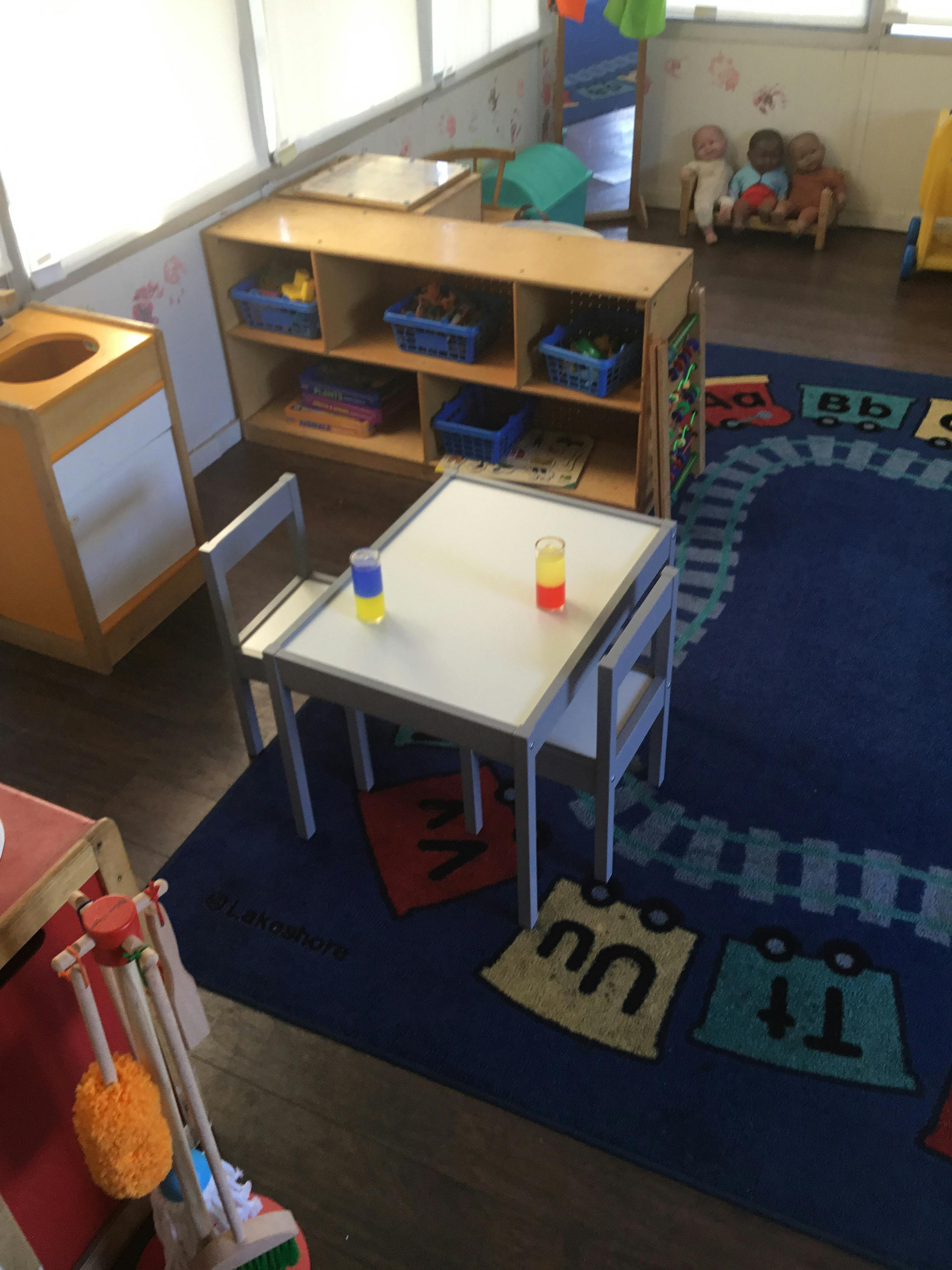 Children's classroom with small table and chairs, shelves with toys, and alphabet rug