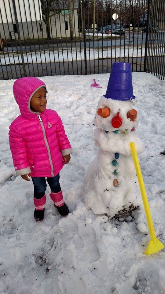 Child in a pink coat standing next to a snowman with a blue bucket on its head