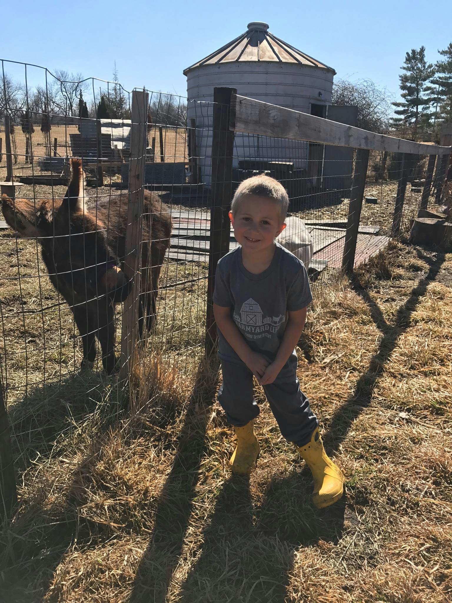 Child in yellow boots standing by a fence with a goat and a silo in the background