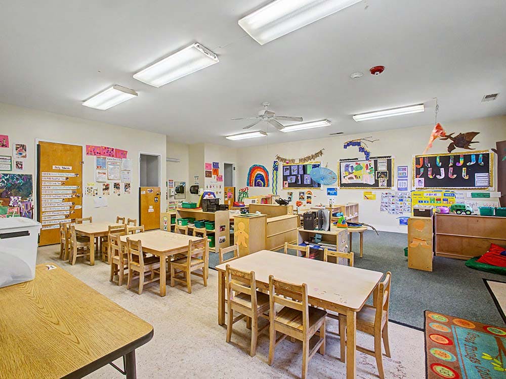 An empty classroom with multiple small wooden tables and chairs, and decorated walls