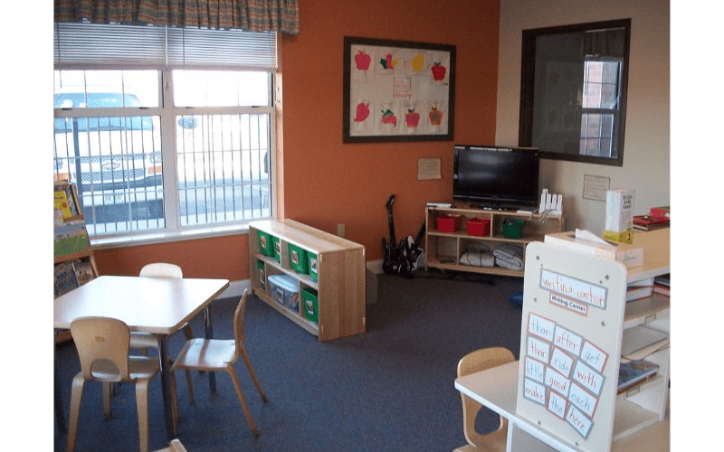 Classroom with small tables, chairs, shelving units, a television, and a writing center
