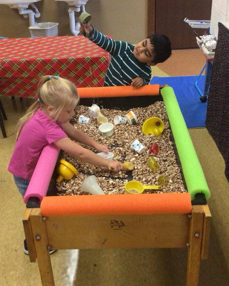 Two children playing at a sensory table with small rocks and cups