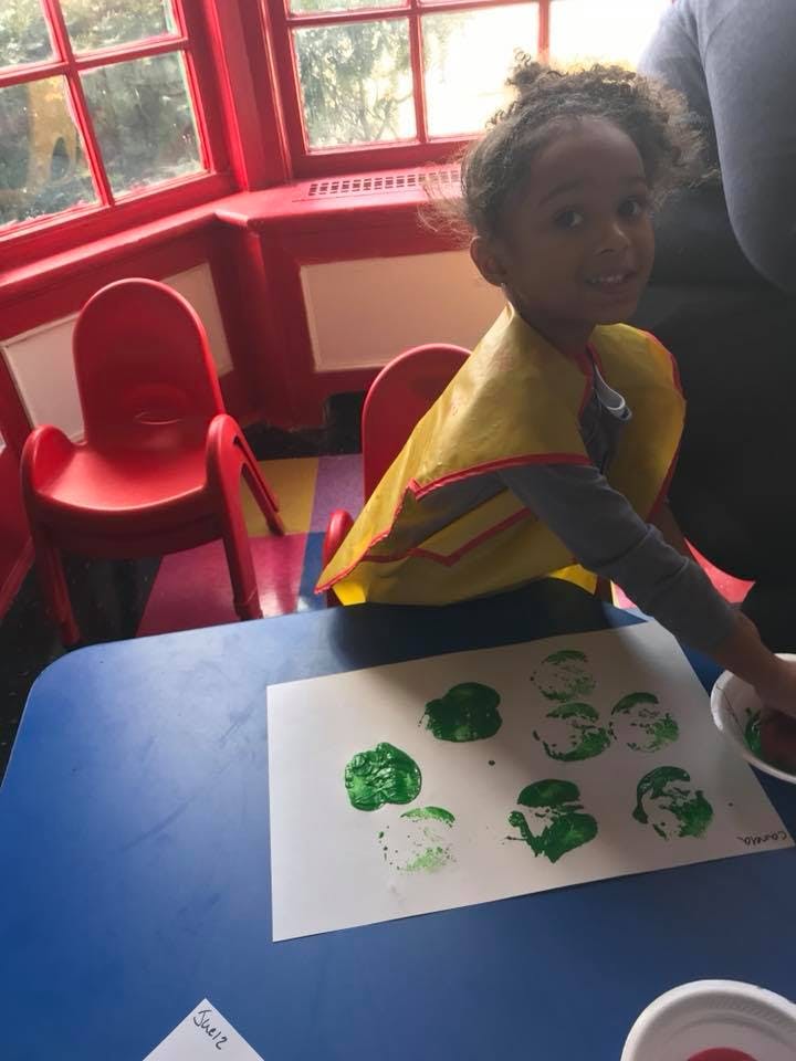 Child in a yellow smock making green prints on paper at a blue table