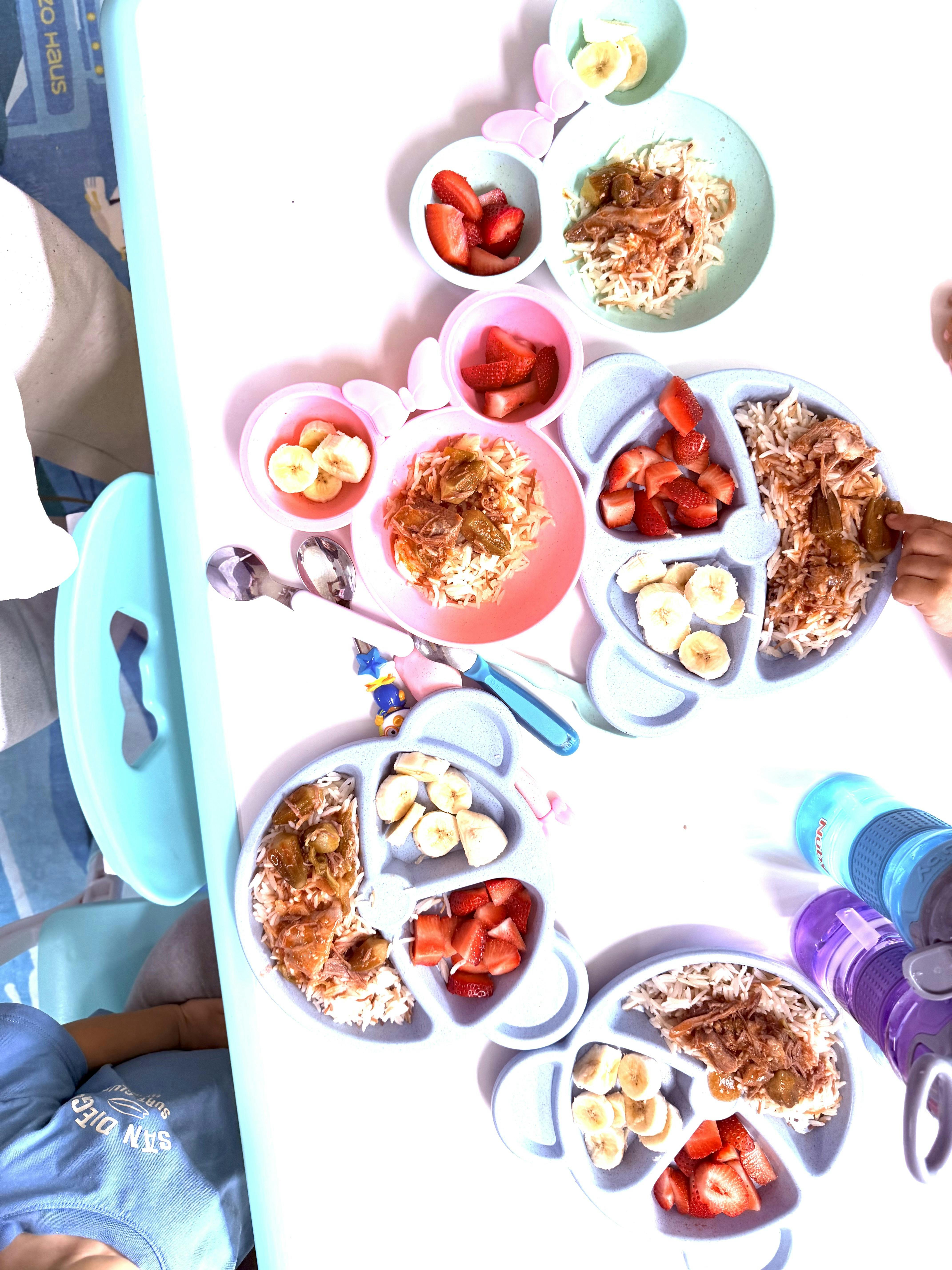 Several divided plates of rice, meat, fruit on a table with a person's hand reaching for food