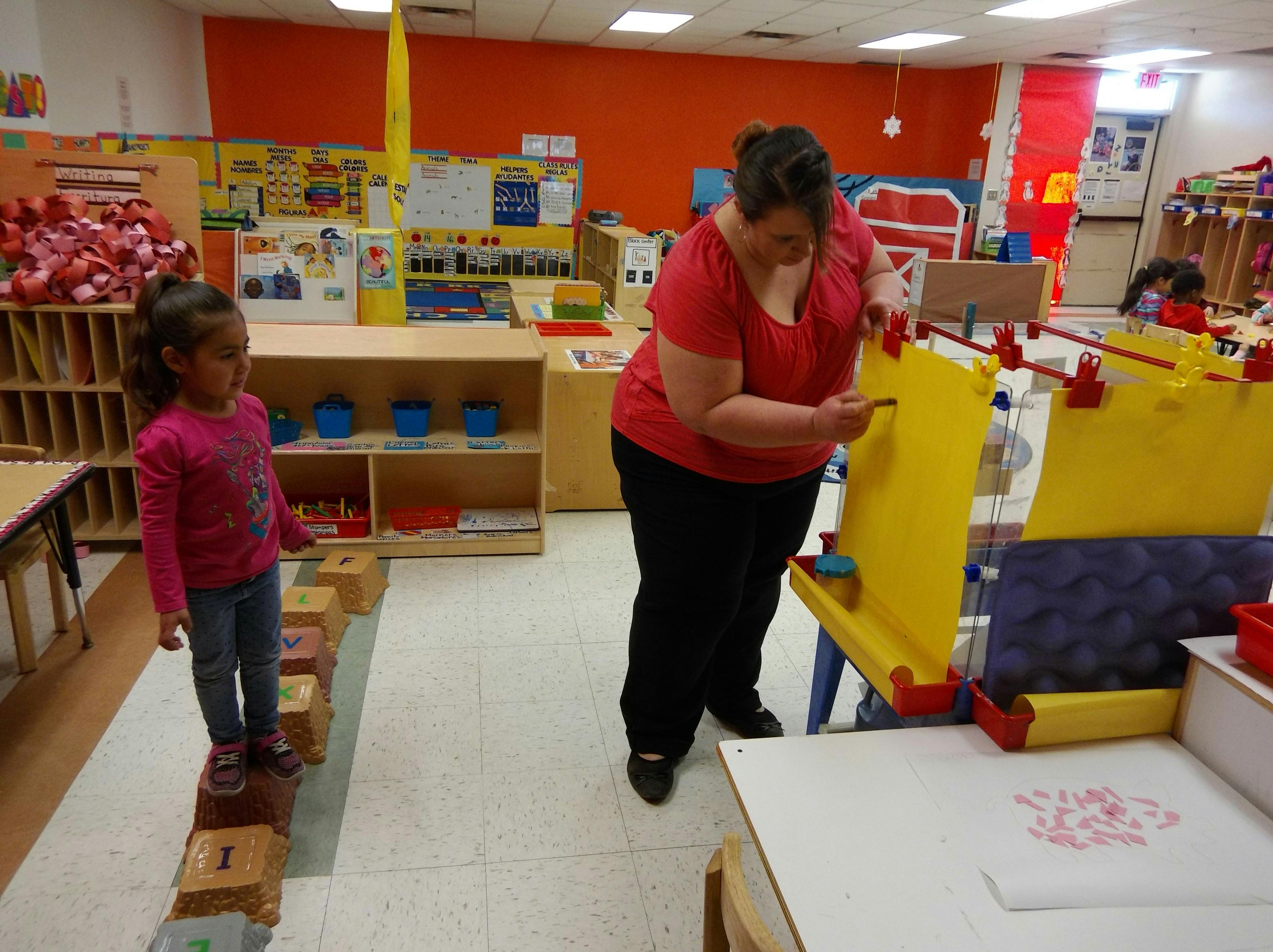 Person drawing on an easel while a child stands on blocks in a classroom