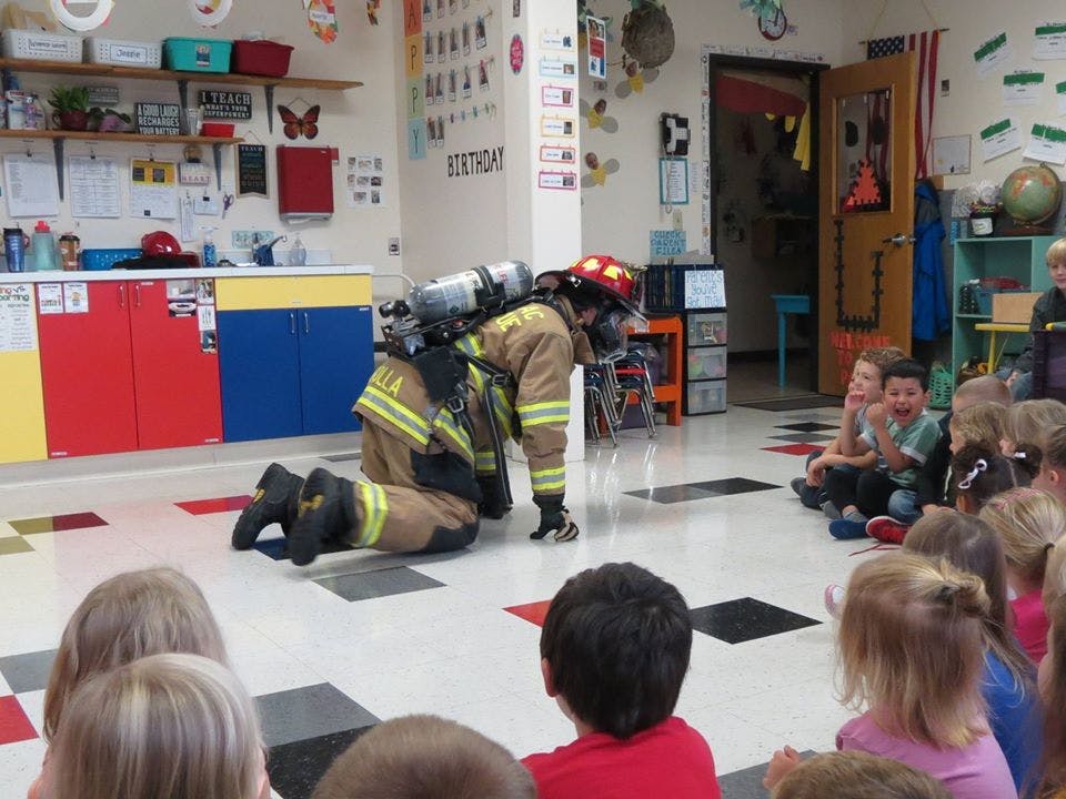 Firefighter in full gear crawling on floor in front of seated children in classroom