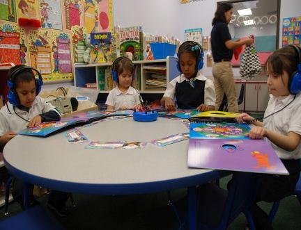 Children wearing headphones reading books at a table in a classroom