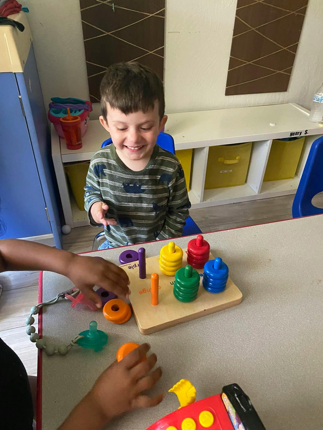 Child smiling while playing with stacking toys at a table with another child