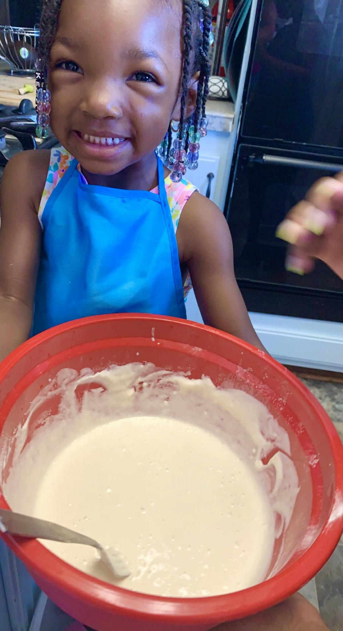 Child wearing an apron holding a large bowl of batter in a kitchen