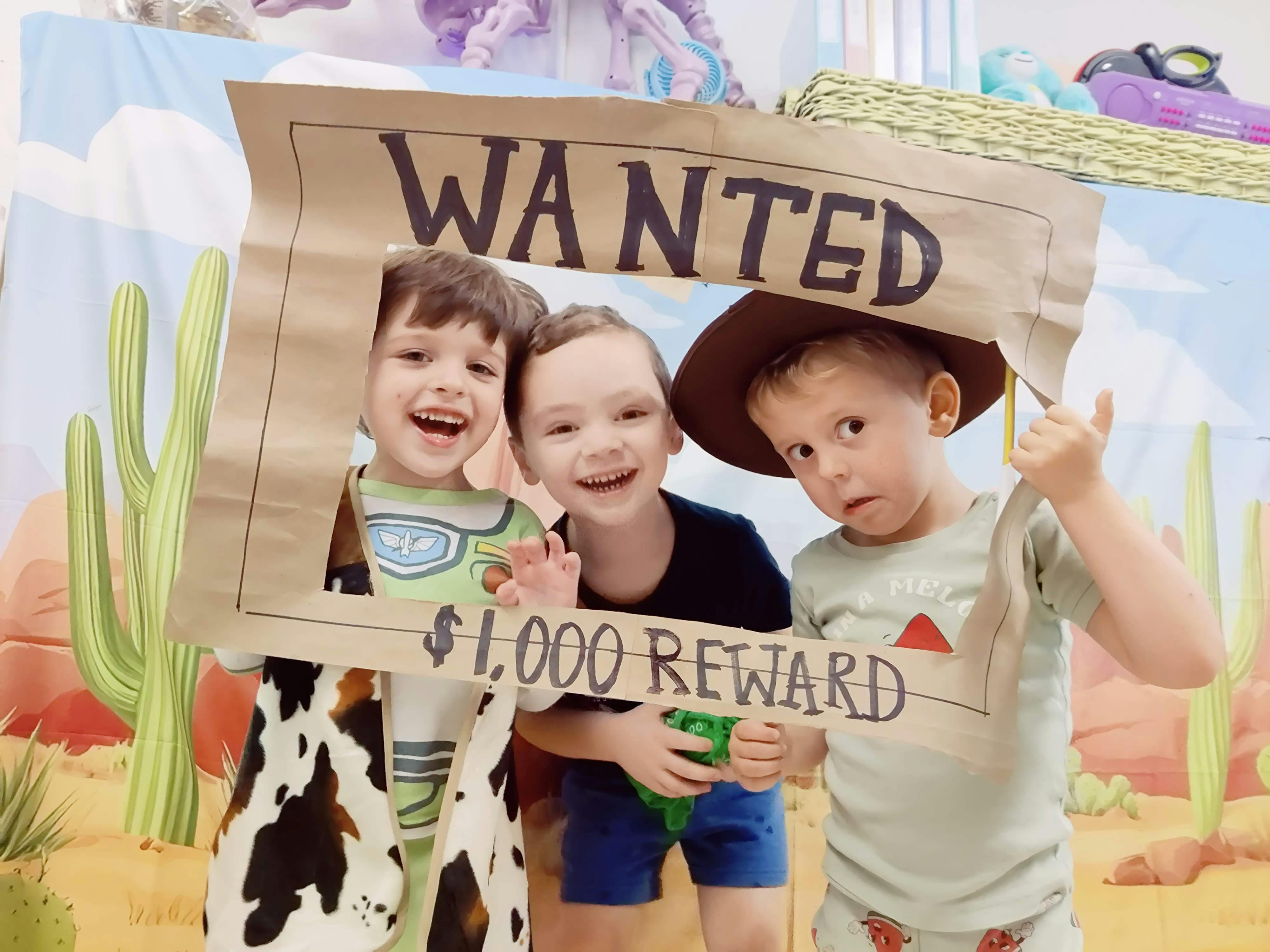 Three children posing behind a cardboard WANTED poster frame