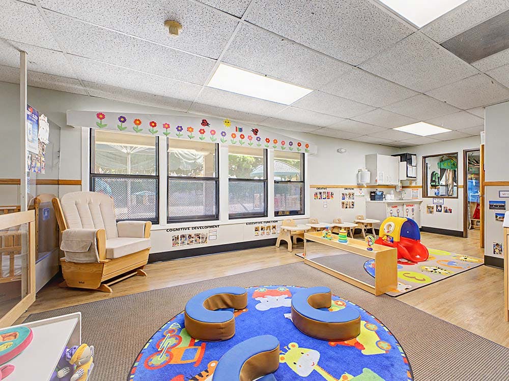 A colorful classroom with a rocking chair, small tables and chairs, and play mats
