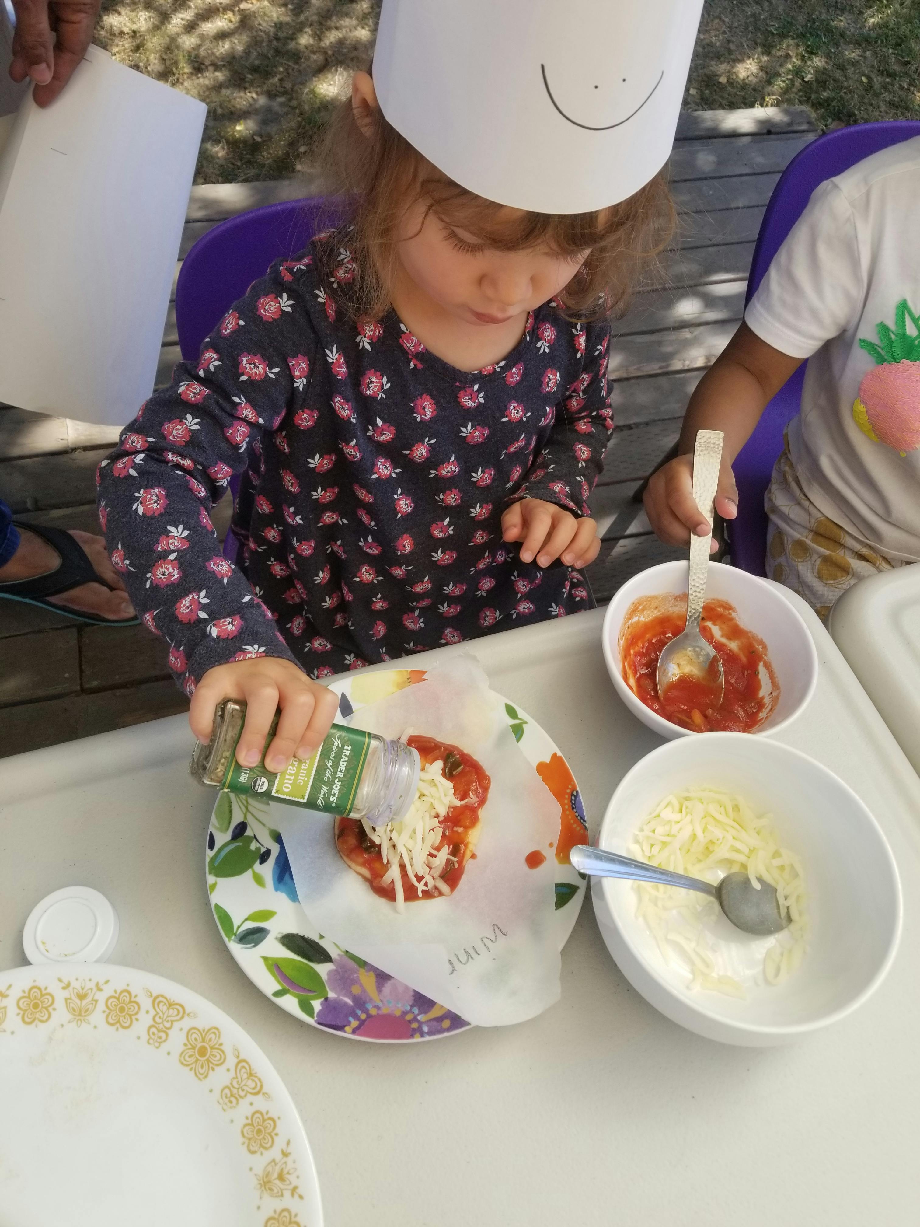 Child in chef hat sprinkling seasoning on a mini pizza