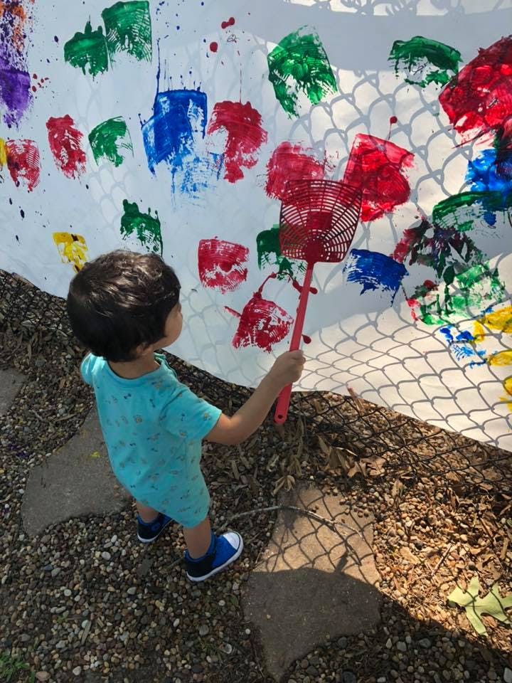 Child holding a fly swatter to a white surface with colorful paint marks