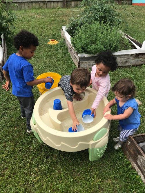 Four children playing with water and cups in a water table outdoors