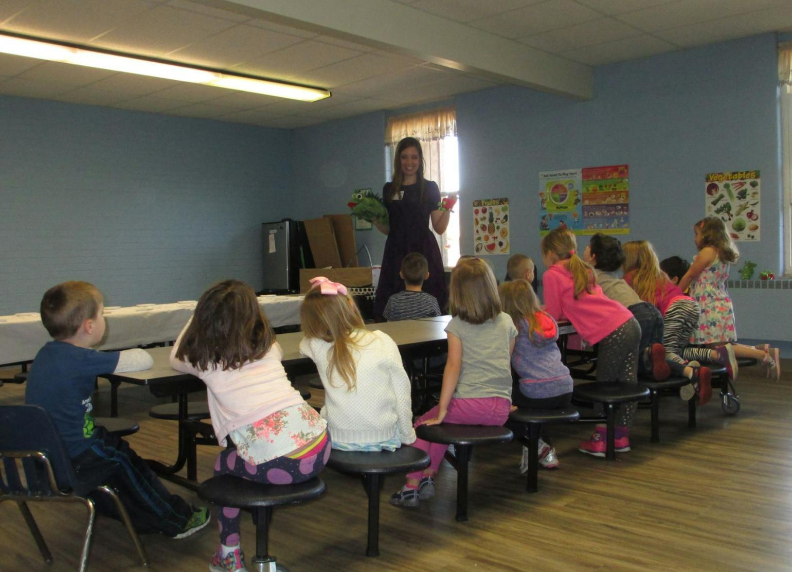 Adult speaking to a group of children in a classroom setting with tables and benches