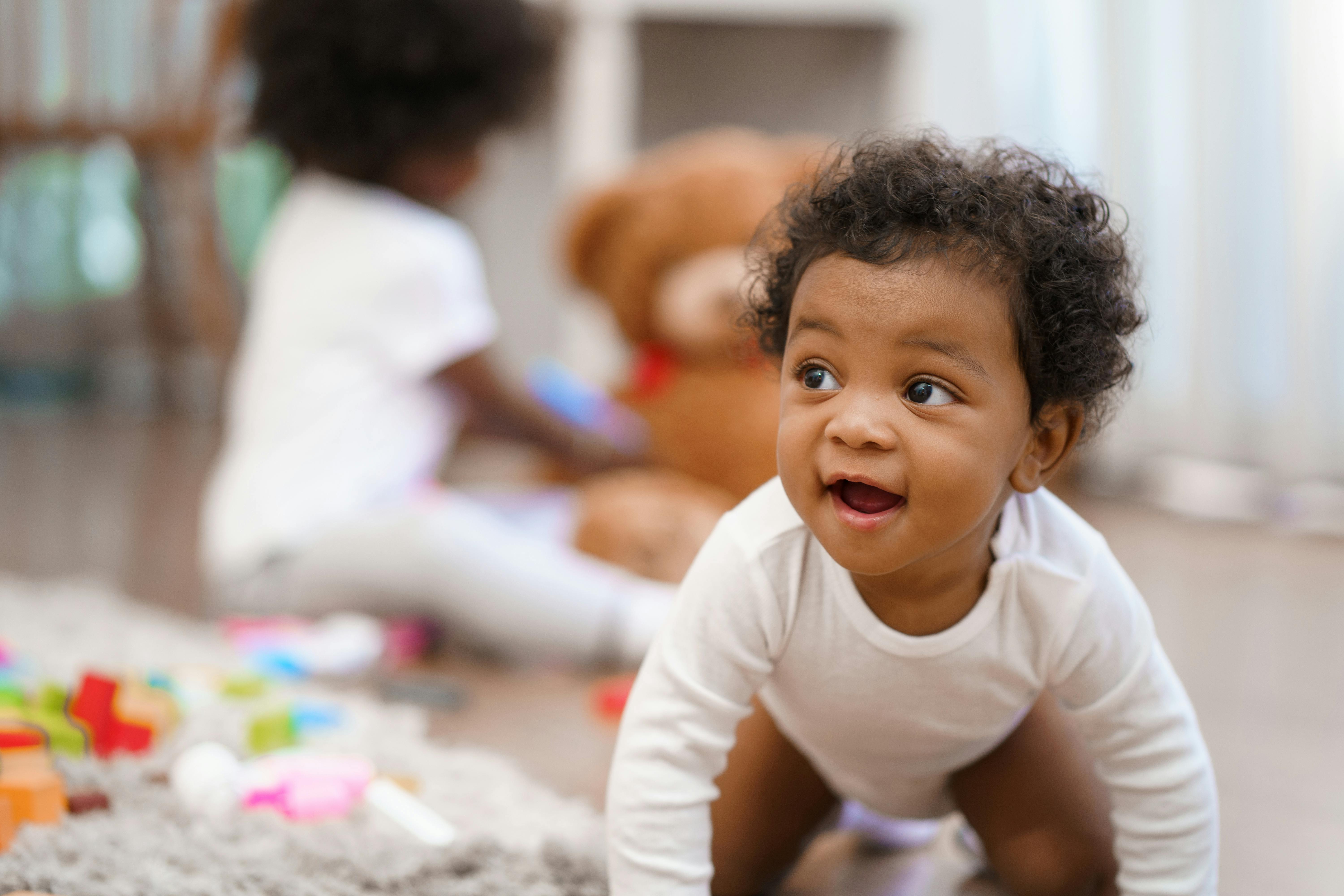 Baby crawling on a rug with another child and toys in the background