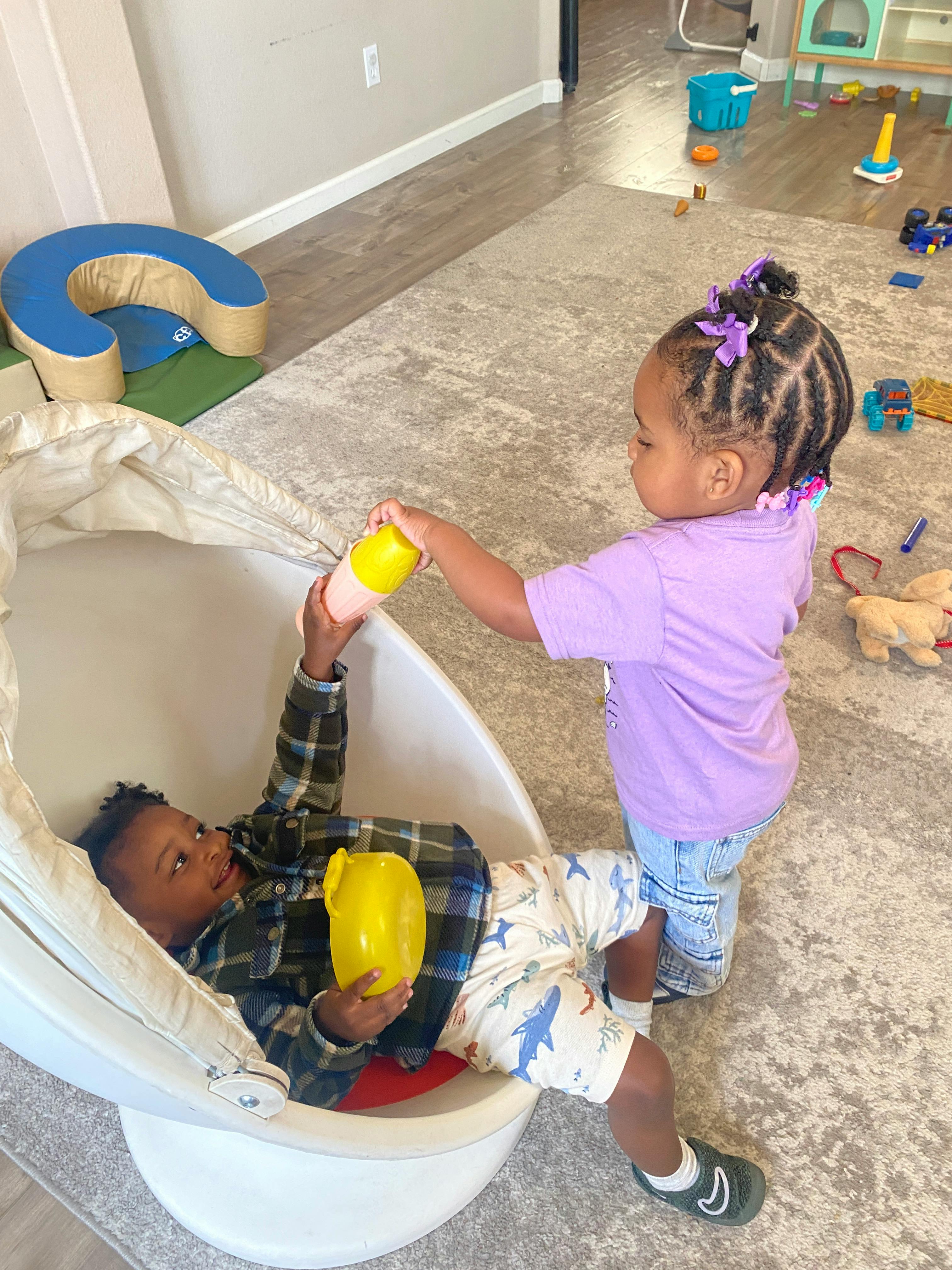 Child in chair holding yellow bottle, another child offering a bottle