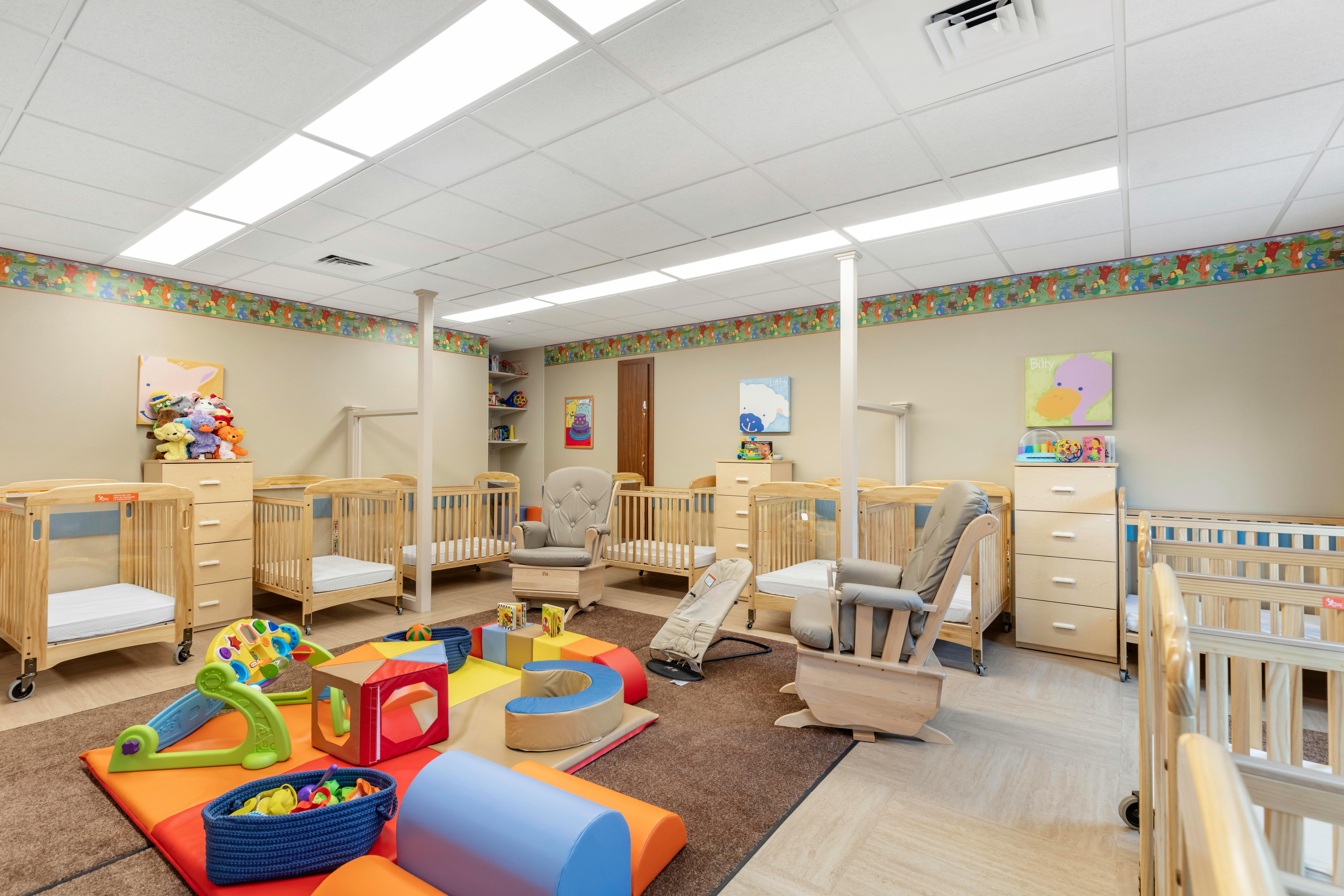 A brightly lit nursery room with multiple cribs, rockers, and soft play mats