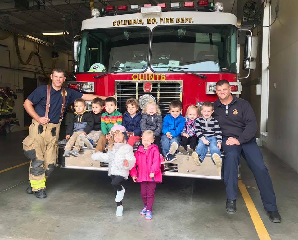 Two people in firefighter uniforms with a group of children on a fire truck