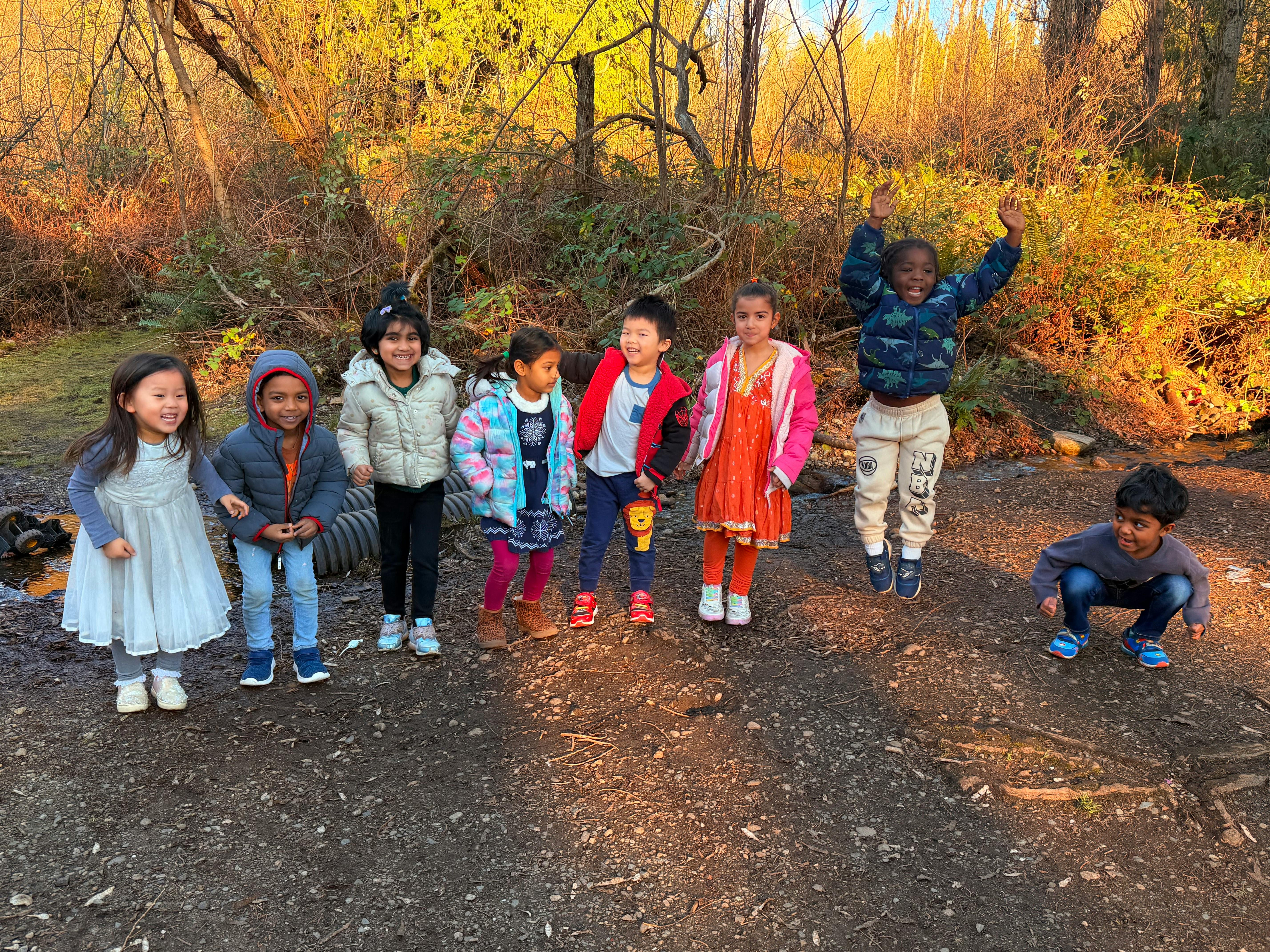 Seven children outside, one jumping, one crouching, others standing and smiling