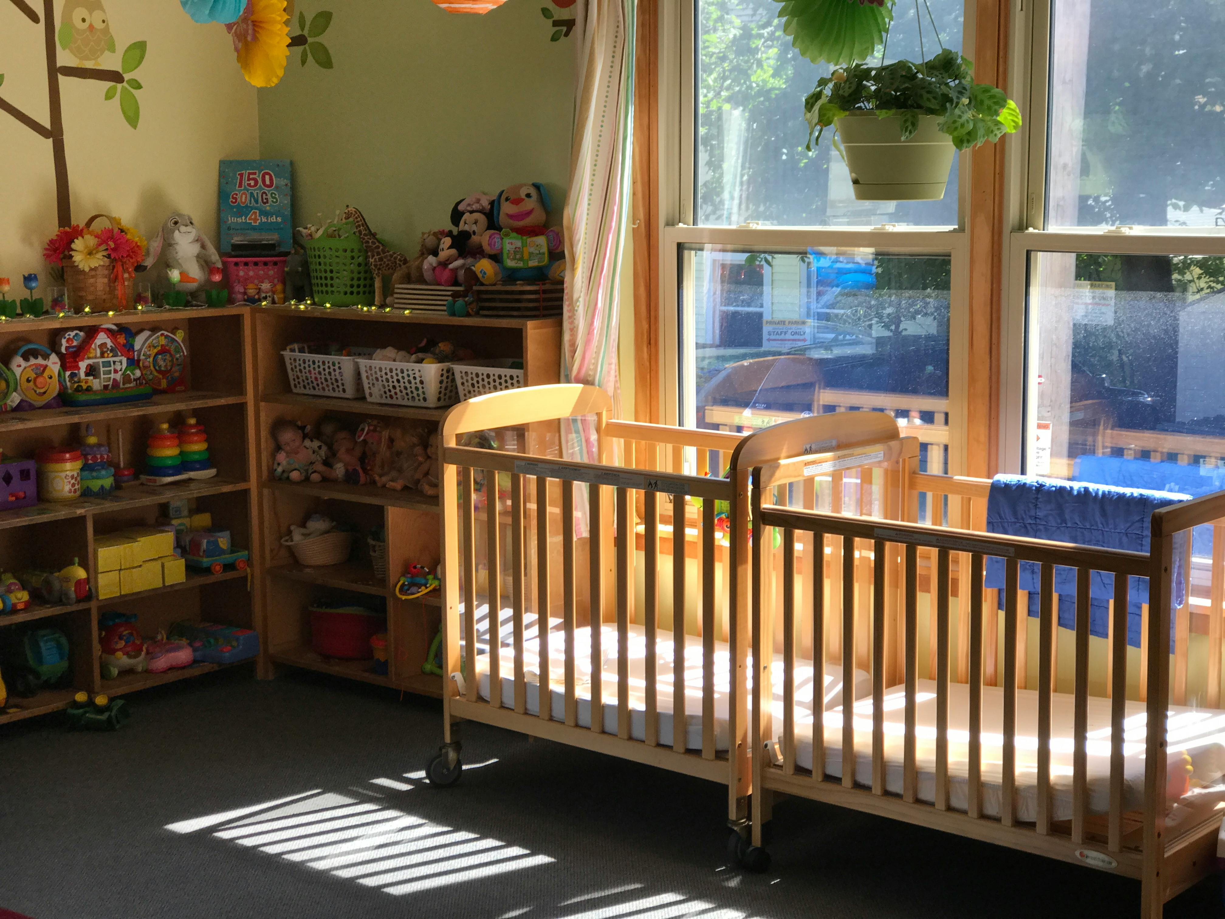 Two empty cribs in a room with shelves of toys and a large window
