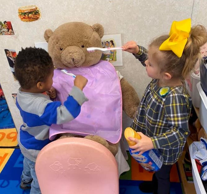 Two children brushing the teeth of a large teddy bear in a classroom