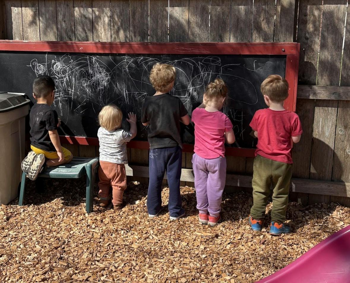 Five children drawing on an outdoor chalkboard mounted to a wooden fence