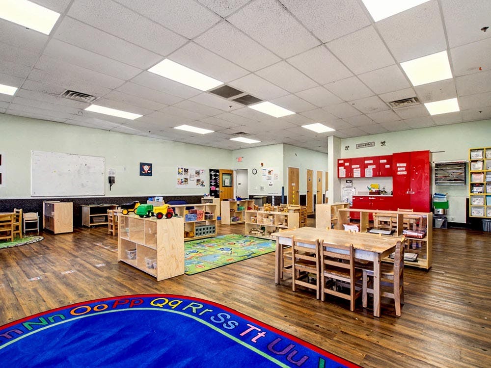 Empty classroom with small tables and chairs, shelves with toys, and an alphabet rug