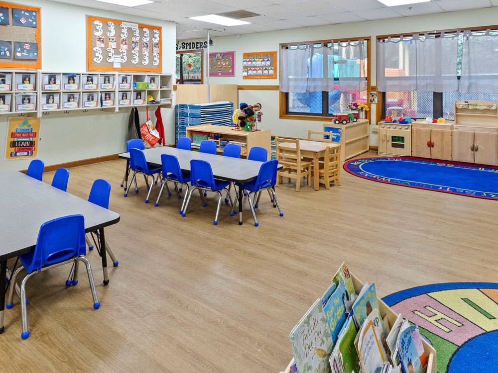 Classroom with small tables and blue chairs, cubbies, play kitchen, and rugs
