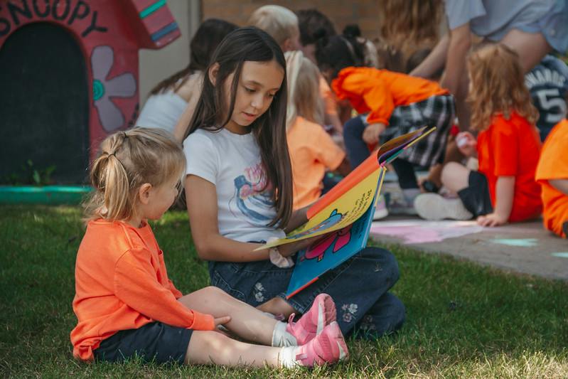 Child reading a book to another child while sitting on the grass