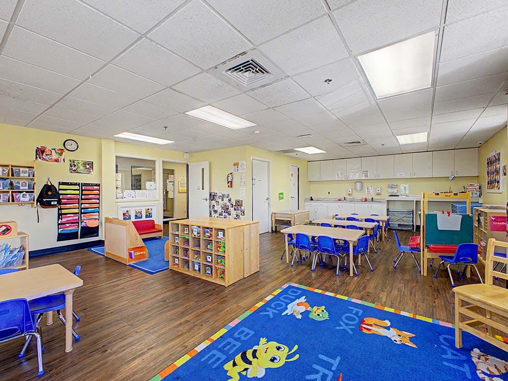 A classroom with small tables and chairs, a play rug, and storage shelves